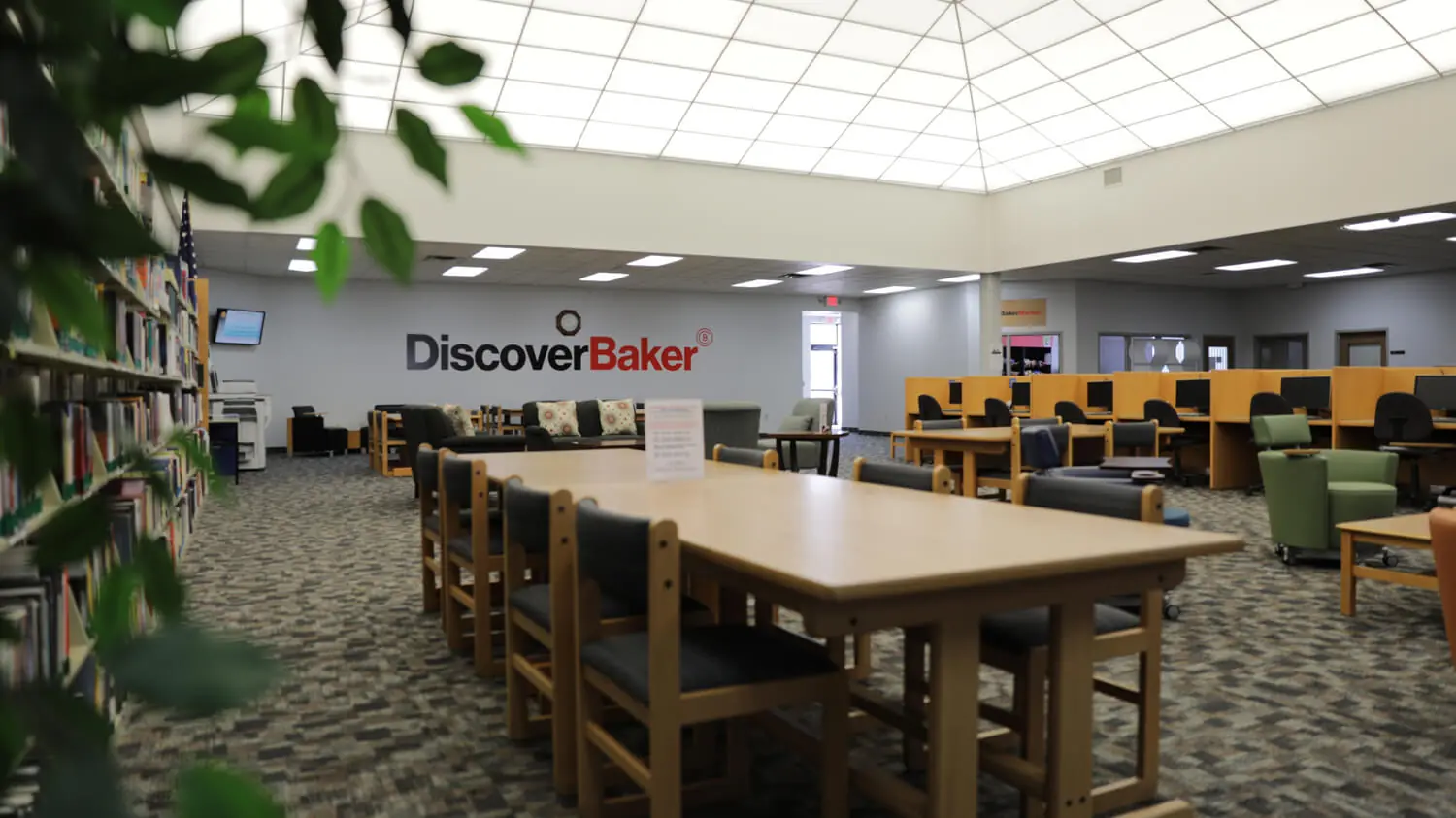 A photo of the Owosso Academic Resource Center with long study tables in the center, a wall of books to the left, and individual computer workstations to the right. The back wall has a large logo that says Discover Baker.