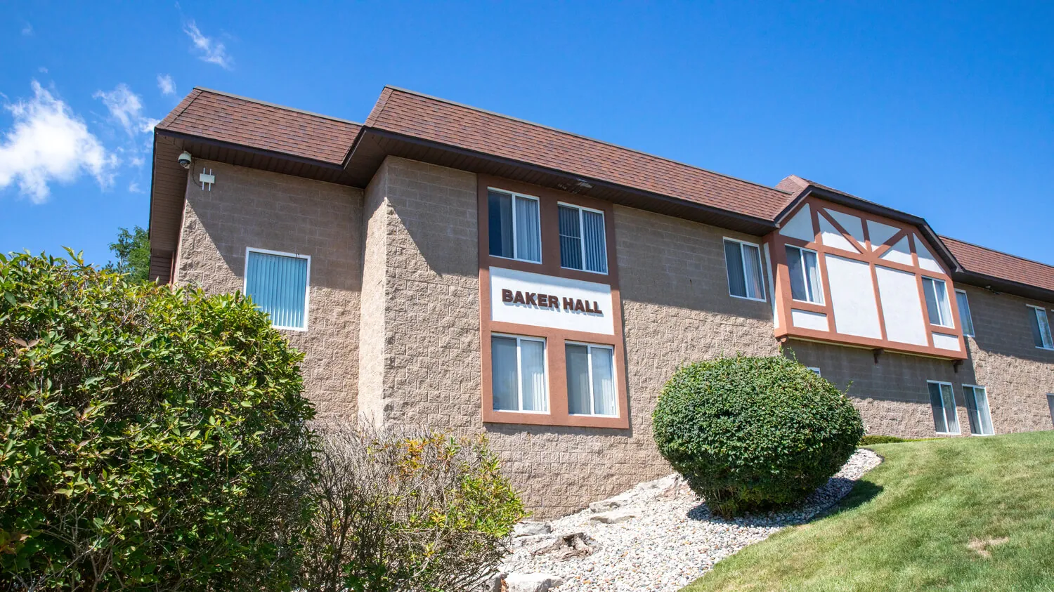 An exterior photo of Baker Hall, a student residence hall on the Owosso campus, showing a cottage-style building on a hillside surrounded by green plants and shrubs. The sky is bright blue and sunny.
