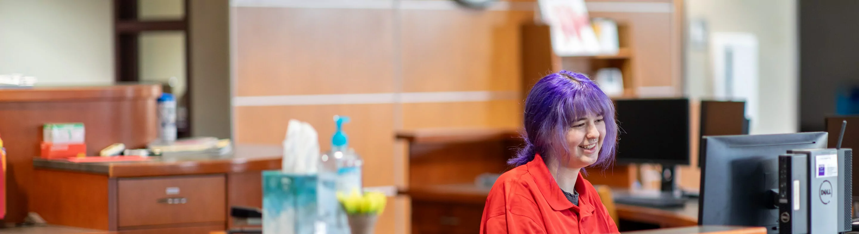 A person in a red shirt sits at a desk in the middle of an office.