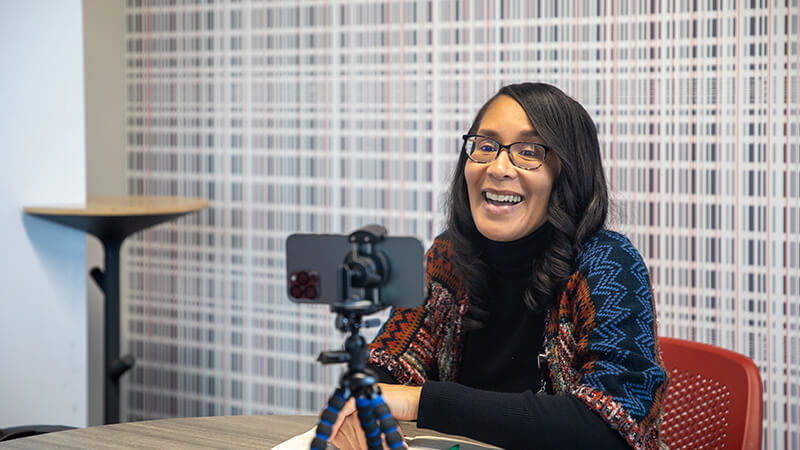 A woman speaks to her phone camera, which is mounted on a tripod.