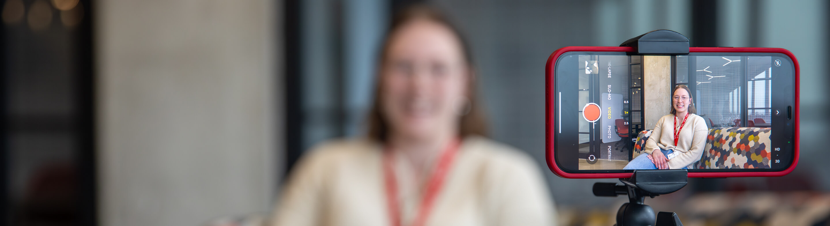 A woman with a red Baker College lanyard as seen through her phone camera, which is mounted on a tripod.
