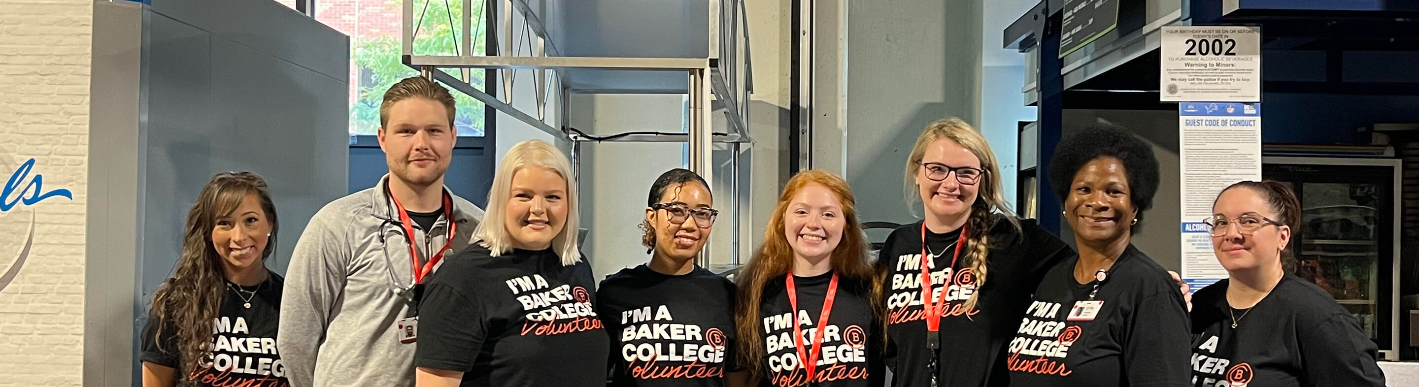 A smiling group of people wearing Baker College Volunteer T-shirts and red lanyards.