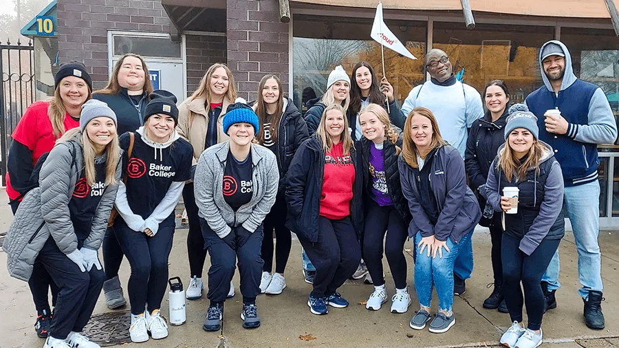 A group of students poses for a photo outside of a campus building. A person in the back holds a white BakerProud flag.