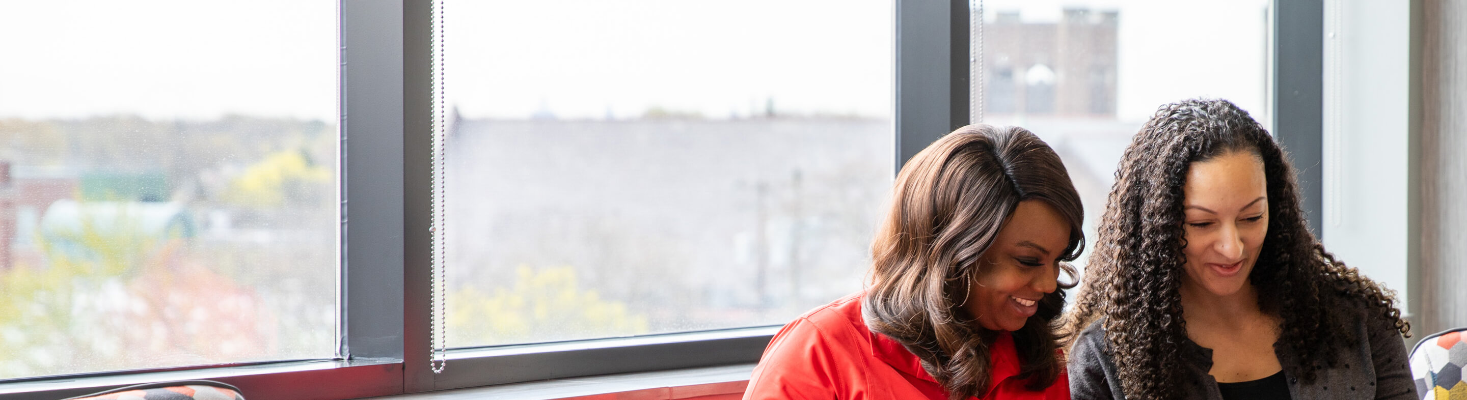Two women review information on a laptop screen together sitting beside a glass window on campus. 