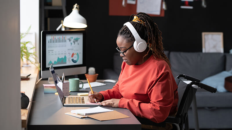 A student takes notes in a notebook with her laptop open while wearing headphones. She's seated at her home desk in a wheelchair.