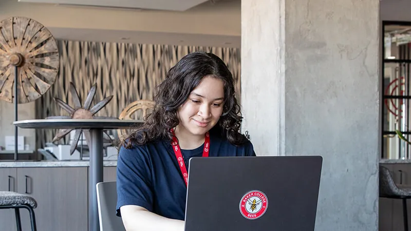 A student wearing a Baker College lanyard works on a laptop with a Baker College Spirit sticker on the cover.