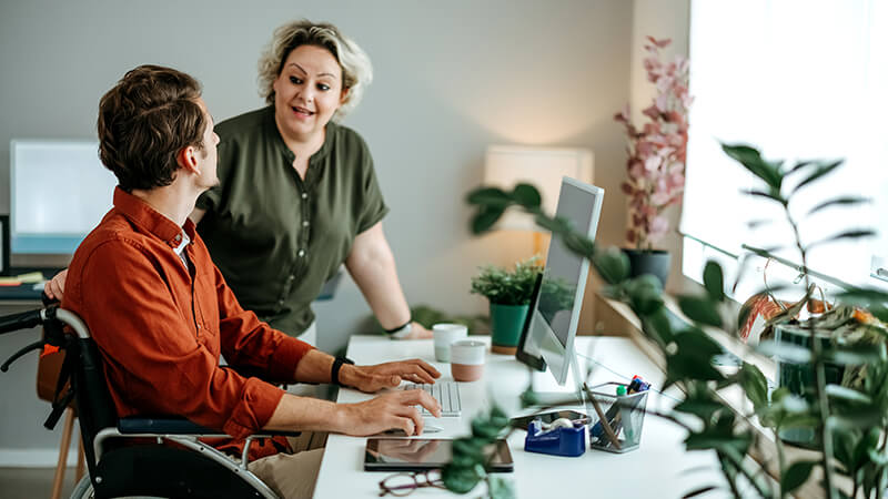 A student sits in a wheelchair in front of a computer and looks up to address a professor standing nearby.
