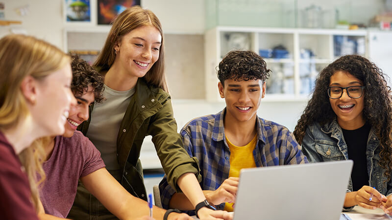 A group of students smile and work together on a project. One student stands and uses a laptop while the rest watch the screen.