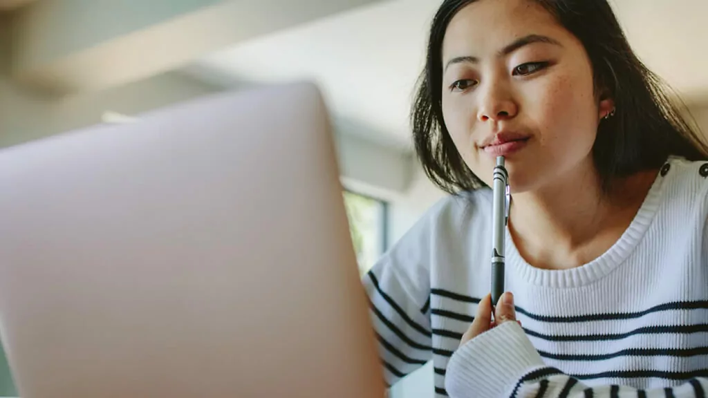 A student looks at her laptop while holding a pen and thinking.