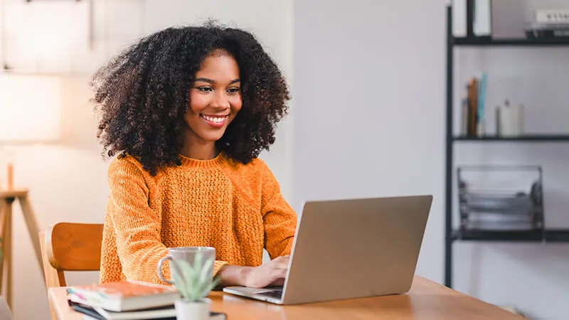 A student smiles as she applies to Baker College on her laptop. She wears an orange sweater and sits in her home office.