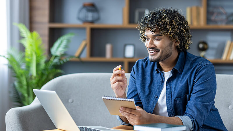 A young man holds a notebook and pencil as he watches his laptop screen while sitting on a sofa in his living room.