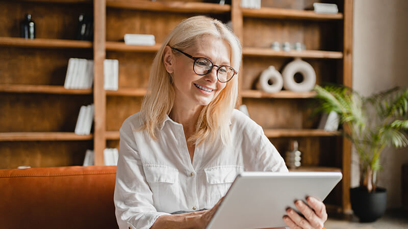 An older woman smiles while holding a tablet while sitting in her living room.