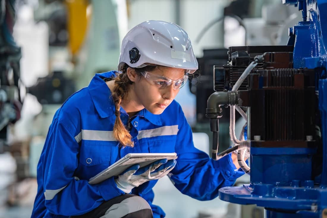 Woman engineer in uniform and helmet doing an inspection check on heavy machine robot arm. 